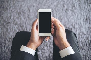 Mockup image of a business man sitting and holding white mobile phone with blank black screen with gray rug in background