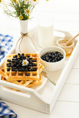 Healthy breakfast: Belgian waffles with blueberries, honey and milk decorated chamomile flowers on white wooden salver. Warm light. Selective focus
