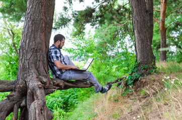 Young man relaxes with laptop sitting in the park, working and relaxing. Freelancer works in the nature, a copy of space.