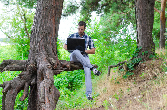 Young man relaxes with laptop sitting in the park, working and relaxing. Freelancer works in the nature, a copy of space.