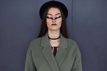 Beautiful, fashionable girl with an unusual makeup in a stylish green coat and black hat in the...