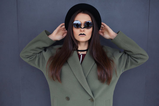 Beautiful, fashionable girl with an unusual makeup in a stylish green coat and black hat in the background of a gray wall.