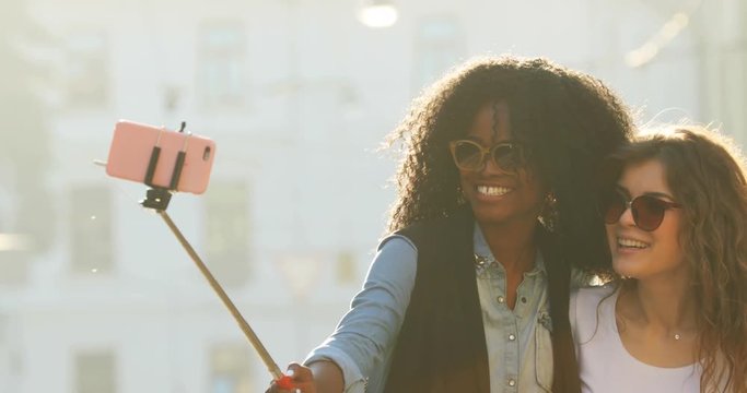 Gorgeous smiling girlfriends with sunglasses are taking photos using the selfie stick while walking the street.