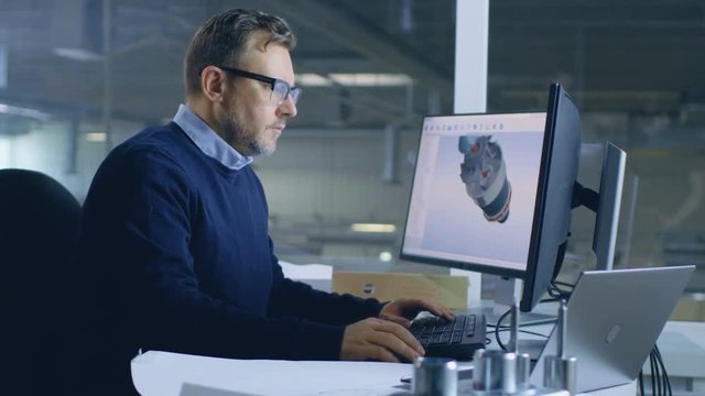 Senior Male Engineer Works on 3D Turbine/ Engine Design on His Personal Computer with Help of Cad Software. In the Background We See Big Factory. Shot on RED EPIC-W 8K Helium Cinema Camera.