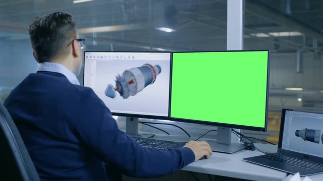 Chief Male Engineer Designs 3D Turbine/ Engine For a Big Industrial Company, His Second Display Shows Mock-up Green Screen Computer. Out of the Office Window Big Factory is Seen. 4K UHD.