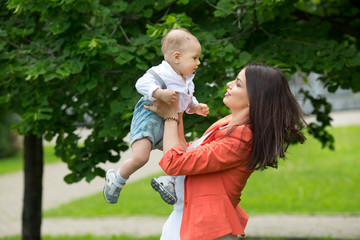 Fototapeta premium A little boy with dark eyes in a white shirt and denim shorts in the hands of his beautiful happy mother in a green park in the summer