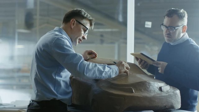 Two Male Automotive Designers Working on a Clay Model of New Generation Electric Car Future Design. One Holds Tablet Computer For Graphic Design, Other Sculpts with Clay with Rake/Wire. 4K UHD.