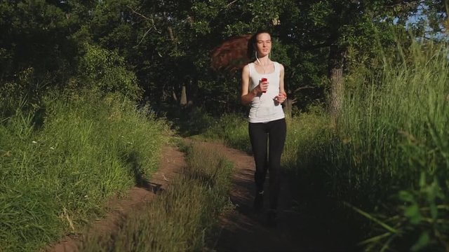 Young sporty woman jogging on the nature. Female listening to the music while running, holding a smartphone on her hands and wearing a smart watches.