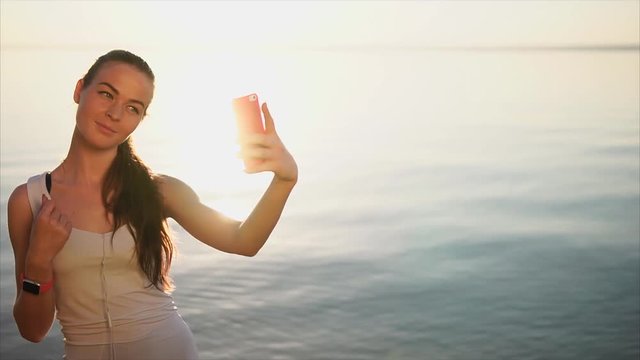 Young pretty woman doing selfie photo using her smartphone. Female standing near the sea during sunset. Enjoyng the nature.