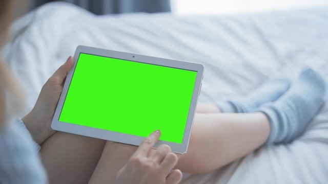 Young Woman in blue sweater sitting on the bed uses Tablet PC with pre-keyed green screen. Few types of gestures - scrolling up and down, tapping, zoom in and out. Perfect for screen compositing