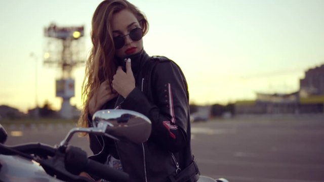 Portrait of a female biker in leather jacket and shorts sitting on her bike and holding a handlebar. Close up of young sexy curly woman in sunglasses on the chopper