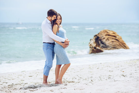 Courageous and handsome man in a white shirt and blue trousers  hugs her beautiful pregnant wife in a blue dress against the background of the rocks and the sea.