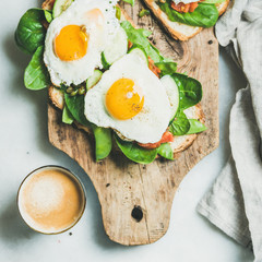 Healthy breakfast sandwiches and cup of coffee. Bread toasts with fried eggs and fresh vegetables on rustic wooden board over grey marble background, top view, square crop. Clean eating food concept