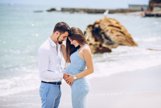 Loving couple on the beach touching pregnant woman's belly with love and care on the background of rocks and sea in sunny day.