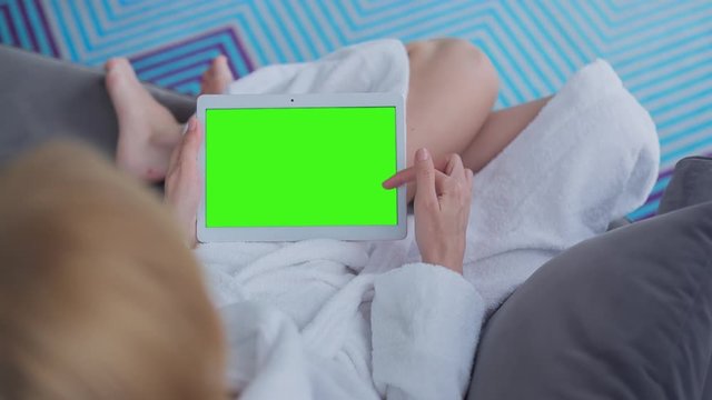 Young Woman in white bathrobe sitting on sofa uses Tablet PC with pre-keyed green screen. Few types of gestures - scrolling up and down, tapping, zoom in and out. Perfect for screen compositing
