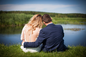 Wedding photography in nature. Bride and groom by the lake. Empty copy space outdoor photoshoot. Young couple love background. Flower crown on brides head. People sitting together in sunlight.