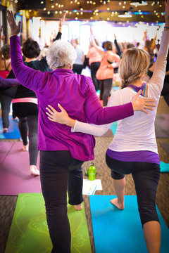 Vertical photo of grandmother and granddaughter in lunge in group yoga practice with colorful mats and festive lighting. Soft focus background with room for text/copy. 