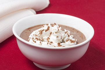 Custard cup of chocolate custard / pudding against a red bckground, topped with whipping cream and powdered cocoa