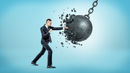 A businessman in full height punching and breaking a huge wrecking ball on blue background.