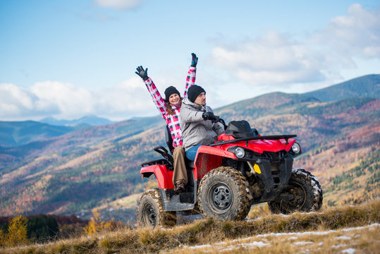 Fototapeta Guy drive atv quad bike, girl sitting behind him and raised her hands up on a mountain road on a background of beautiful landscape mountains and blue sky