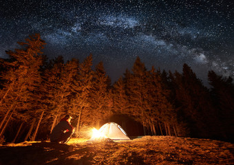 Male tourist have a rest in his camp near the forest at night. Guy sitting near campfire and tent under beautiful night sky full of stars and milky way. Long exposure