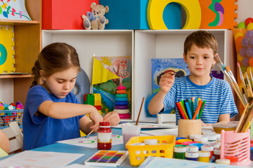 Small students girl and boy painting in art school class. Child drawing by paints on table. Craft drawing education develops creative abilities of children. Toy bear on shelves background.