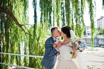 Newlywed standing in the park under the willow and holding champagne glasses with a lake in the background.