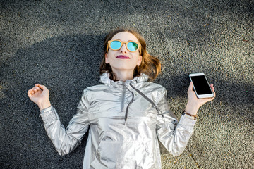 Lifestyle portrait of a modern woman in silver jacket lying with phone outdoors on the asphalt road. Smart phone with empty screen to copy paste