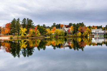 Obraz premium Buildings Among Colourful Trees and Reflection in Water ion a Cloudy Autumn Day. Lake Placid, NY.