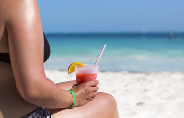 Tanned woman in bikinis sitting on the beach and holding a red cocktail with straw on her lap.