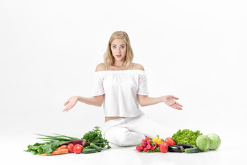 Frustrated blond woman spreads hands in both directions and lots of fresh vegetables on white background