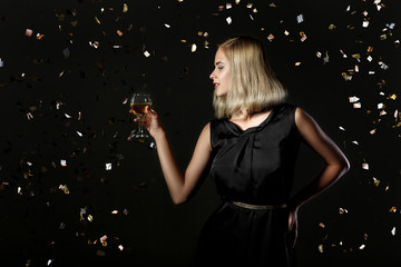 Beautiful happy blonde woman holding glass of white wine on black background with confetti