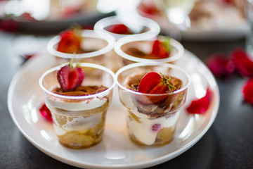 Delicious fresh cold strawberry cheese cake, dessert in plastic cups with rose petals on a dark background. Copy space