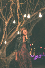 Woman among decorative outdoor string lights hanging on tree in the park at night time. Bali island, Indonesia.