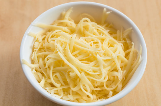Close-up of Grated Gouda cheese in white cup on wood table.