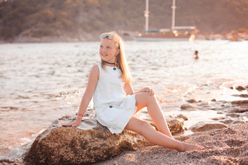 Romantic and happy blonde child cute girl sit on the stone near water at the beach in sunset magic light. Stylish Kid has fun on the sea coast. Summer vacation and active lifestyle concept