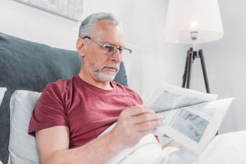 focused senior man reading newspaper while lying in bed at home