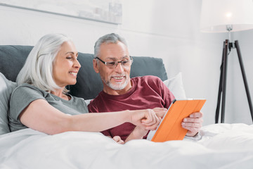 happy senior couple lying in bed and using tablet together