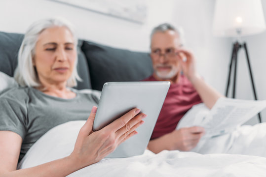selective focus of woman using tablet while husband reading newspaper in bed