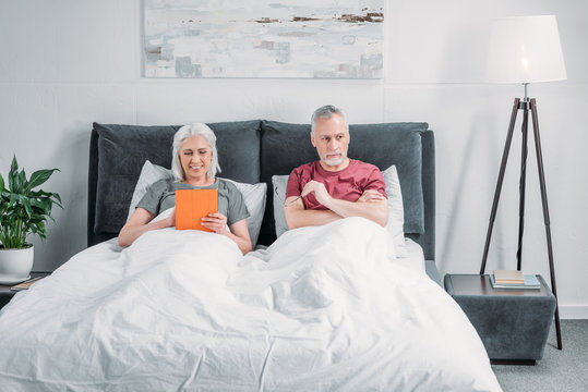 woman using tablet while pensive husband lying in bed near by