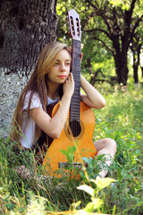 The lovely girl sits thoughtfully, leaning on a guitar against the background of nature..
