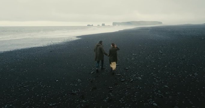 Back aerial view of the young couple walking on the black volcanic beach in Iceland. Romantic date near the sea.