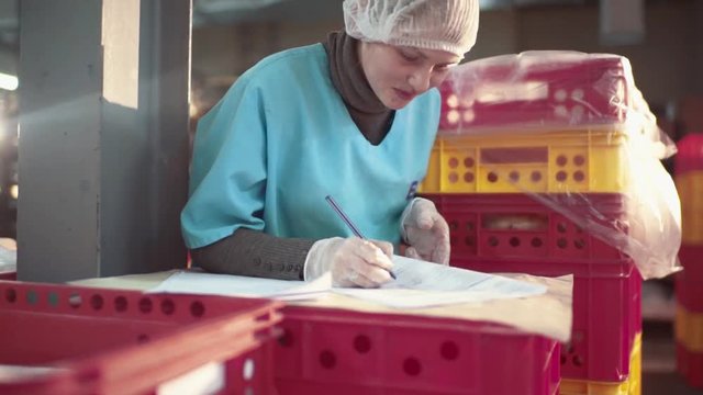 Rotation view of young smiling female bakery-worker filling in the papers, surrounded with trays with bread. Transporting the production, bread manufactory. Healthy food, natural ingredients.