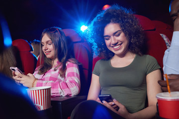 Young cheerful women smiling using their smart phones while sitting at the cinema auditorium watching a movie technology mobility connection communication friendship youth entertainment activity.