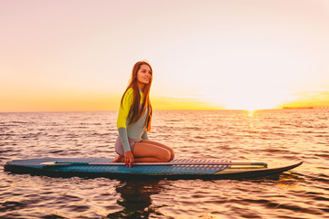 Stand up paddle boarding on a quiet sea with warm sunset colors. Happy young woman relaxing on ocean