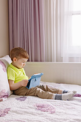 Portrait of cute adorable white Caucasian toddler boy sitting in bed playing with digital tablet with funny face expression. Candid lifestyle early development. New technology generation.