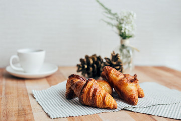 Side view of fresh crispy butter croissants laying on wood cutting board with flower and pine decoration and white cup of black coffee, beautiful layout perfect for breakfast