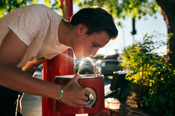 Cute handsome american young hipster teenager man in white tshirt drinks water from street fountain, with beautiful shadow sun light leaks beaming on drops, healthy hydration