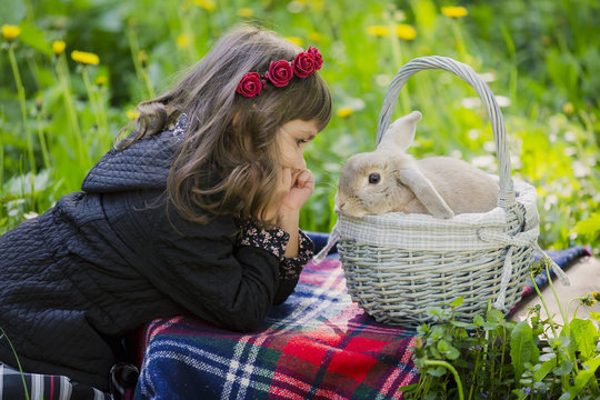A little girl in a wreath observes a rabbit in a basket at sunset in a park.