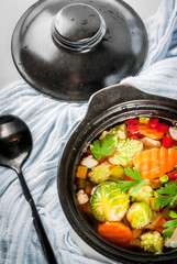Healthy eating. Dietary vegan dinner. Homemade soup with fresh spring and summer vegetables cabbage, broccoli, green peas, carrots, celery. In black serving pan, white marble table. Copy space